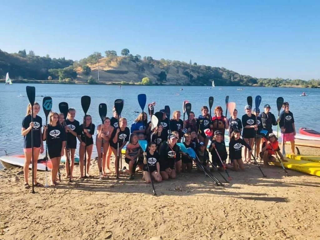 Youth race team posing on a lakefront beach with paddles raised